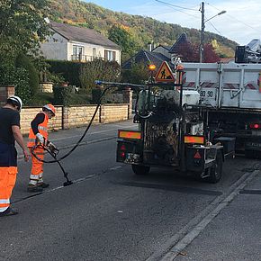 Deux ouvriers travaillent à la refection de la voirie derrière un camion transportant le mélange de bitume et d'elastomère 